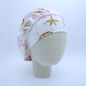 May include: White scrub cap with a pink and gold sea-themed pattern. The pattern includes starfish, seashells, and seahorses. The cap is designed to fit comfortably and securely, ideal for medical professionals or anyone needing hair protection.