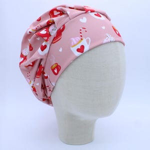 May include: A pink beanie hat with a repeating pattern of red and white hearts, coffee cups, and cats. The hat is made of a soft fabric and has a knotted top. The hat is displayed on a mannequin head.