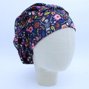May include: A dark blue surgical scrub cap with a floral and skeleton print. The cap features a repeating pattern of colorful flowers and white skeletons. The cap is displayed on a mannequin head.