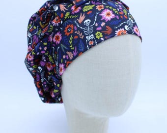 Könnte beinhalten: Eine dunkelblaue OP-Haube mit Blumen- und Skelettmuster. Die Haube hat ein sich wiederholendes Muster aus bunten Blumen und weißen Skeletten. Die Haube wird auf einem Schaufensterkopf präsentiert.