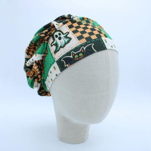 May include: A slouchy beanie hat featuring a festive Halloween print with white ghosts, black bats, and orange and black checkered patterns on a green and white background. The fabric has a ribbed texture and a gathered design at the crown.