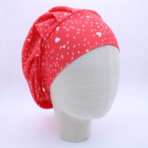 May include: A red fabric hat with a white heart and speckle pattern. The hat is displayed on a mannequin head. The hat has a gathered design with a bow detail on the side. The hat is made of a soft, lightweight material.