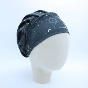 May include: A dark charcoal gray scrub cap featuring a celestial print with white and pale yellow suns, crescent moons, and scattered stars. The cap has a gathered, pleated design at the crown and a wide, fitted band around the forehead area.