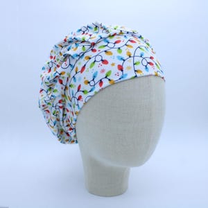 May include: A white bouffant surgical scrub cap with a colorful string of Christmas lights pattern. The cap features red, green, blue, and yellow lights and small star accents. The cap is displayed on a mannequin head.