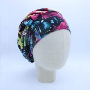 May include: A colorful, patterned beanie featuring a butterfly design. The hat displays a vibrant mix of pink, blue, yellow, and black hues. The beanie is displayed on a mannequin head.