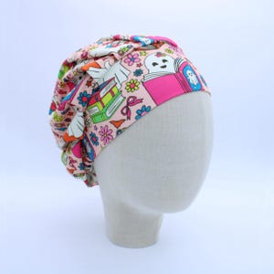 May include: A pink scrub cap featuring a repeating pattern of white ghosts, colorful books, flowers, and small bats. The fabric has a ribbed texture and is gathered at the top, displaying a vibrant print with bright blue, green, and yellow accents.