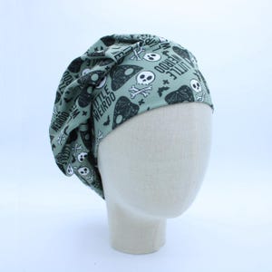 May include: A sage green scrub cap featuring a repeating pattern of black and white skulls, crossbones, bats, and ouija board planchets. The fabric is printed with the text LITTLE WEIRDO in a bold, black sans-serif font throughout the design.