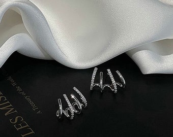 Puede incluir: Un par de pendientes tipo ear cuff plateados con un diseño de garra de cuatro puntas. Cada punta está decorada con pequeñas piedras claras y brillantes en el borde exterior. Las piezas de joyería están sobre una superficie oscura junto a tela blanca.