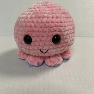 May include: A round, light pink crochet plush toy shaped like an octopus. It features two large, glossy black safety eyes and a small, stitched black mouth. The underside of the tentacles shows a contrasting light purple colour.