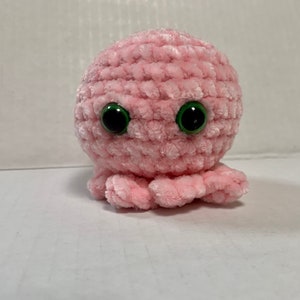 May include: A round, light pink crocheted plush toy featuring a soft, textured yarn surface. The object has two large, prominent, glossy black eyes with green irises positioned on the front, and several small, curled tentacles at the base.