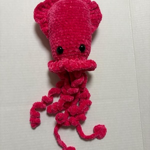 May include: A handmade, plush, magenta crochet jellyfish. The toy features a rounded head with ruffled edges, two small black eyes, and long, curly tentacles. The tentacles end in heart-shaped tips. The jellyfish is made of soft, velvety yarn.