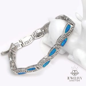 May include: Silver-toned bracelet with blue opal accents. The bracelet features a Greek key design and oval-shaped blue opal stones. The Jewelry Junction logo is visible in the bottom right corner.