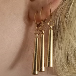 May include: Three gold dangle earrings with a long, thin, cylindrical shape. The earrings are hanging from a person's ear.