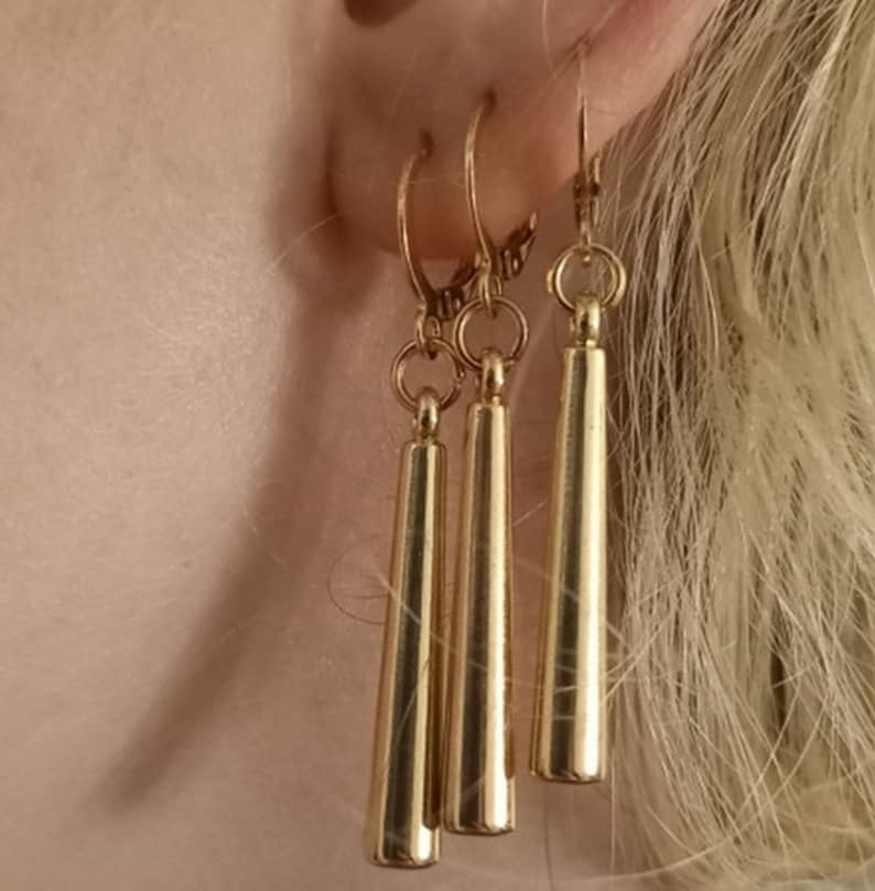 May include: Three gold dangle earrings with a long, thin, cylindrical shape. The earrings are hanging from a person's ear.
