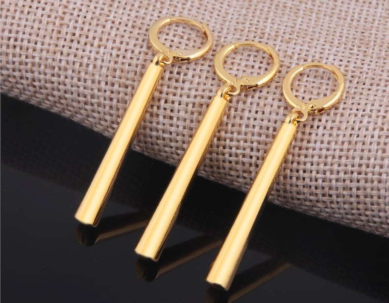 May include: Three gold-colored dangle earrings with a long, thin, cylindrical design. The earrings have a hoop closure.