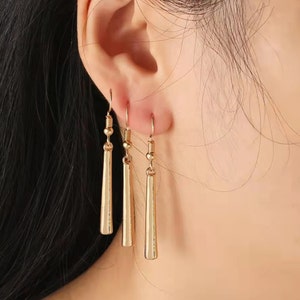 May include: A pair of gold dangle earrings with a long, thin, straight design. The earrings are made of metal and have a simple, elegant style.