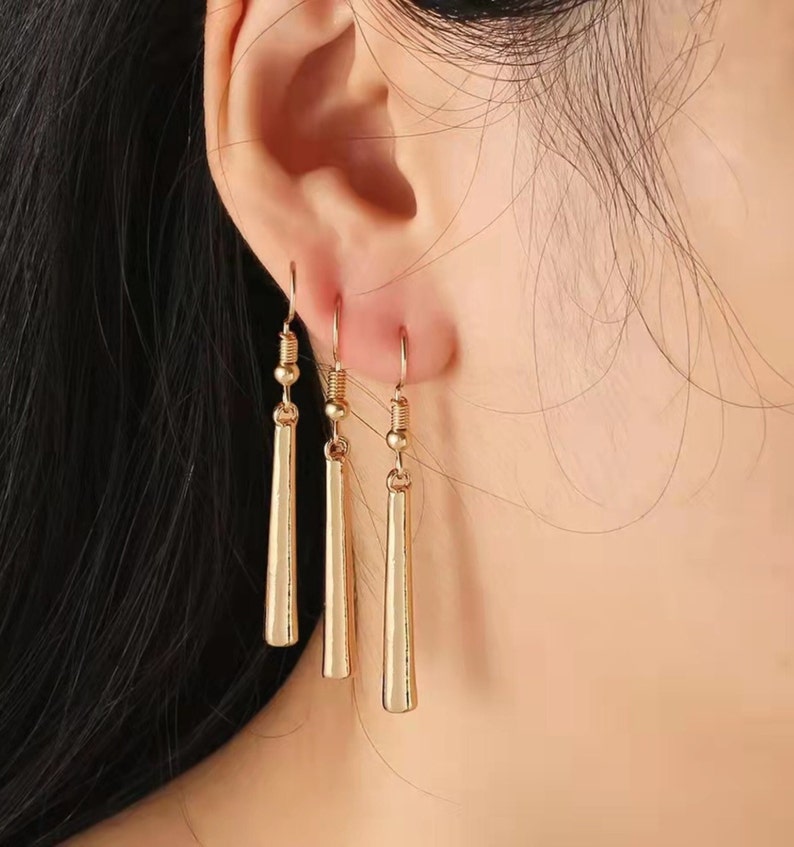 May include: A pair of gold dangle earrings with a long, thin, straight design. The earrings are made of metal and have a simple, elegant style.