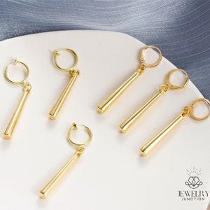 May include: Six gold-colored clip-on earrings with long, thin, straight, dangling charms. The earrings are on a white surface.