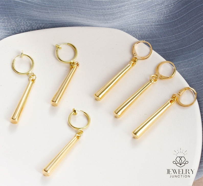 May include: Six gold-colored clip-on earrings with long, thin, straight, dangling charms. The earrings are on a white surface.