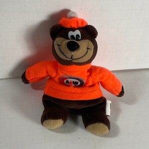 Pode incluir: Um urso de pelúcia marrom vestindo um suéter e chapéu laranja, com o logotipo da A&W no suéter. O urso tem um rosto sorridente e é um item de colecionador.