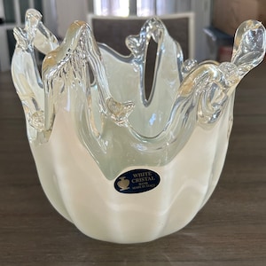 May include: A decorative white and clear glass bowl with a unique, wavy rim design. The bowl has a smooth, opaque white base that transitions to a transparent, textured rim. The label reads "WHITE CRISTAL HAND MADE IN ITALY."