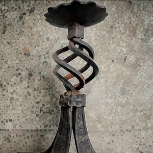 May include: A dark gray metal candle holder featuring a scalloped circular top plate, a central twisted spiral stem, and a base composed of three curved legs with a hammered texture. The metal surface shows signs of oxidation and rust.