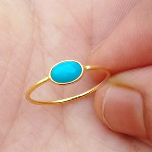Könnte beinhalten: Ein dünner Goldring mit einem kleinen, glatten, oval geformten Türkisstein in einer Zargenfassung. Der Ring hat eine schlichte, polierte Goldschiene, die den Finger umschließt und die leuchtend blaue Farbe des zentralen Steins hervorhebt.