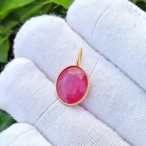 Op de afbeelding: Een gefacetteerde, ovaalvormige robijn hanger gezet in een gepolijste goudkleurige metalen zetting. De hanger is voorzien van een klein goudkleurig oogje aan de bovenzijde voor bevestiging aan een ketting, met een levendige dieproze-rode kleur.
