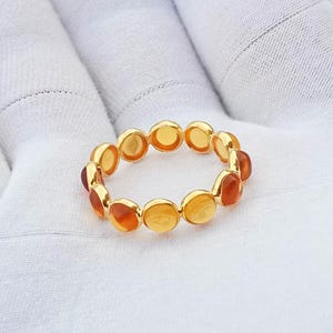 May include: A gold ring featuring a series of small, circular, bezel-set amber stones in varying shades of orange, yellow, and deep reddish-brown. The stones are arranged in a continuous band, creating a linked, circular design with a polished finish.
