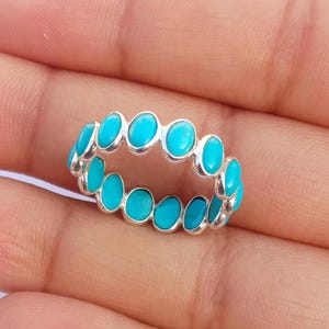 May include: A silver eternity band ring featuring a continuous row of small, oval-shaped, bright turquoise blue stones set in polished metal bezels that encircle the entire circumference of the band, creating a uniform and repeating pattern.