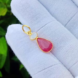 May include: A gold pendant featuring a teardrop-shaped red gemstone suspended below a small, round, translucent white gemstone. The pendant includes a circular gold bail at the top for attachment to a chain, showcasing a polished metal finish.