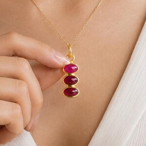 May include: A gold chain necklace featuring a vertical pendant with three stacked, oval-shaped, deep pink cabochon gemstones set in gold bezels. The necklace is worn against a white garment, highlighting the contrast between the gold and pink.