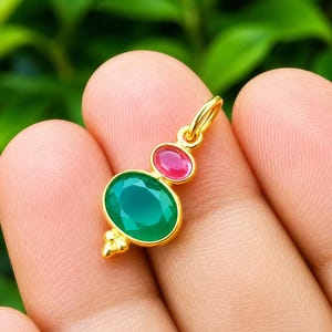 May include: A gold pendant featuring a large oval green gemstone and a smaller oval pink gemstone set in a yellow gold frame. The pendant includes a small gold jump ring at the top for attachment and decorative gold bead accents at the base.