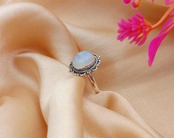 May include: A silver ring featuring a large, oval moonstone with an iridescent sheen. The stone is set in a decorative, filigree bezel. The ring is presented on peach-coloured satin fabric, with pink flowers in the background.
