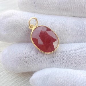 Op de afbeelding: Een gouden hanger met een ovale, gefacetteerde, rode edelsteen. De edelsteen is gezet in een gouden bezel en heeft een kleine gouden lus aan de bovenkant voor een ketting. De hanger wordt gepresenteerd op een wit oppervlak.