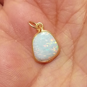 Puede incluir: Un colgante de oro con una piedra de ópalo blanco. El ópalo tiene una forma ligeramente irregular y está engastado en un bisel de oro.