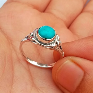 Peut inclure: Une bague en argent ornée d'une pierre turquoise ronde et polie sertie dans un chaton. L'anneau est décoré d'un motif de corde torsadée autour de la pierre et de petites perles d'argent sur les côtés, créant un design détaillé.