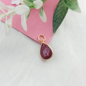 May include: A gold-coloured pendant featuring a teardrop-shaped, deep red gemstone. The pendant has a small loop at the top for a chain. The background includes pink paper and white flowers.