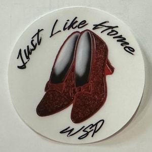 Może przedstawiać: Okrągły biały podkładka z napisem "Just Like Home" napisanym czarnym kursywą. Podkładka przedstawia parę brązowych butów z czerwonym obcasem. Napis "4/SP" jest napisany czarnym kursywą na dole podkładki.