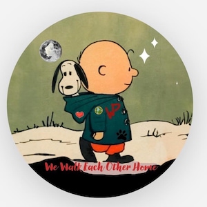 May include: A circular graphic print featuring a cartoon character in a teal hooded jacket with a small dog peeking out from the hood. The character wears orange pants and black boots. The text "We Walk Each Other Home" is printed in red at the bottom.