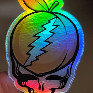 May include: Holographic sticker featuring a peach with a lightning bolt design and a skull. The sticker has a rainbow color scheme and a leaf at the top. The skull has a red and orange color scheme.