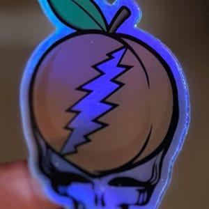May include: A holographic sticker featuring a peach design with a lightning bolt and a skull. The peach has a green leaf and is outlined in black. The sticker has a blue iridescent glow.