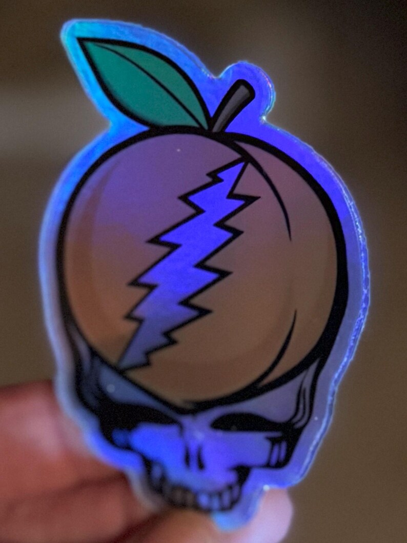 May include: A holographic sticker featuring a peach design with a lightning bolt and a skull. The peach has a green leaf and is outlined in black. The sticker has a blue iridescent glow.