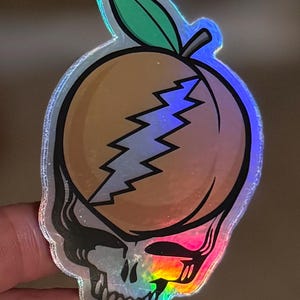 May include: Holographic sticker featuring a peach design with a lightning bolt and a skull. The peach is orange with a green leaf and a silver outline. The skull is black and white with a rainbow gradient.