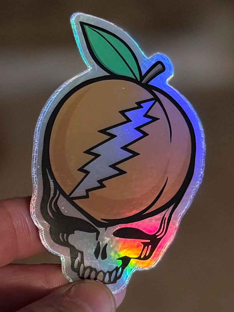 May include: Holographic sticker featuring a peach design with a lightning bolt and a skull. The peach is orange with a green leaf and a silver outline. The skull is black and white with a rainbow gradient.