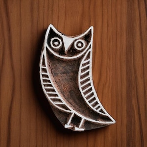 Puede incluir: Un bloque de madera con forma de búho tallado. El búho tiene un cuerpo marrón con detalles blancos, incluyendo los ojos, el pico y los patrones de las alas. El bloque está sobre un fondo de madera marrón.