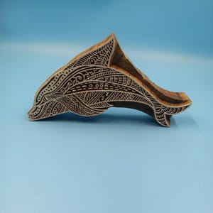 Könnte beinhalten: Ein Holzblock mit einem Delfin-Design. Der Delfin ist mit komplizierten Mustern und Linien geschnitzt. Der Block besteht aus hellbraunem Holz und ist vor einem blauen Hintergrund platziert.