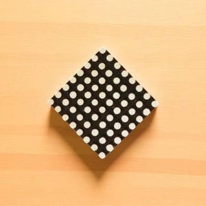 May include: A black and white polka dot square coaster, set diagonally on a light wood surface. The coaster features a repeating pattern of white circles on a black background. The coaster is a square shape.