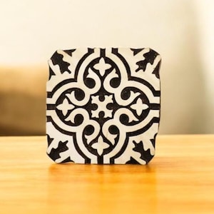 May include: Square, black and white patterned coaster with a symmetrical design. The coaster features a repeating floral and geometric pattern. The coaster is sitting on a wooden surface.