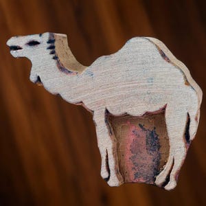 May include: A wooden camel block print, painted white with brown accents. The camel is standing with its head turned to the left. The block print is a traditional craft used for creating patterns on fabric.